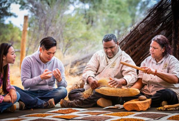 A Karrke Aboriginal Cultural Experience & Tours guide and a woman demonstrate traditional seed grinding to two guests. They sit cross-legged on the ground with various seeds displayed on mats in the Northern Territory Red Centre. © Tourism Australia