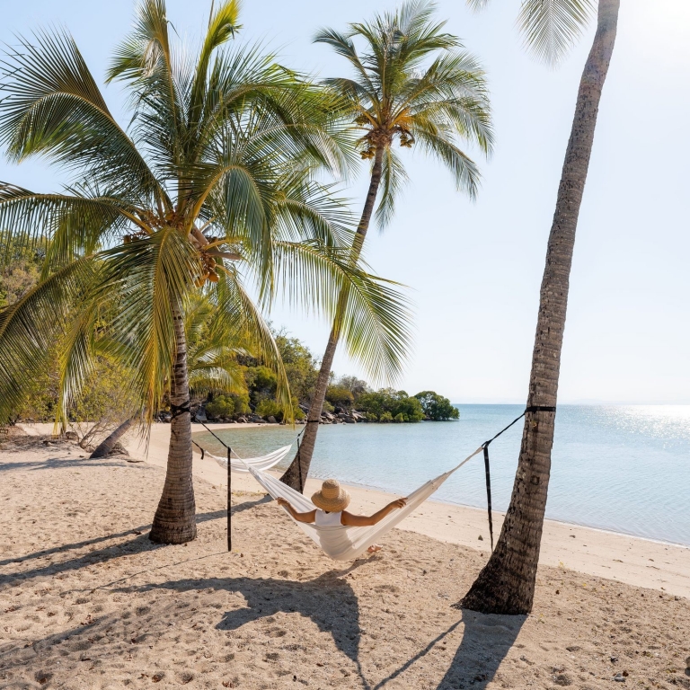 A person in a wide-brimmed sunhat relaxes on a hammock at Orpheus Island Resort, gently swaying between two tall palm trees. The golden sand beneath them leads to a calm, crystal-clear bay that glistens in the sunlight. The scene is peaceful and tropical, with the palm trees casting soft, dappled shadows. © Tourism Australia