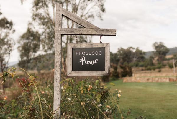A wooden sign reading "Prosecco Picnic" hangs from a timber post amidst rose bushes on a lush vineyard lawn at Pizzini Wines, High Country, Victoria. © Tourism Australia