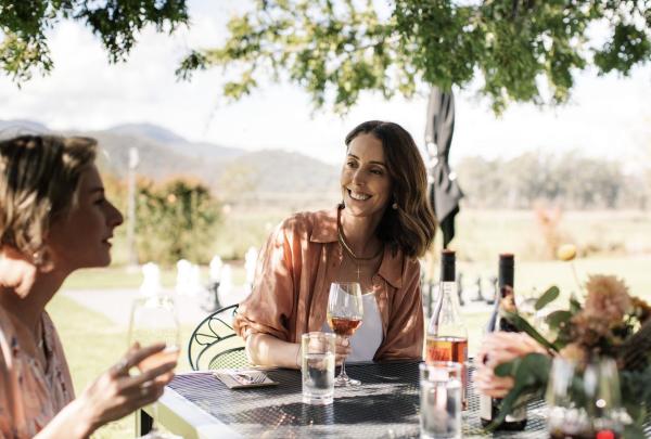 Two women sit outside enjoying glasses of wine at a table under leafy trees with mountain views in the background at Pizzini Wines, High Country, Victoria. © Tourism Australia