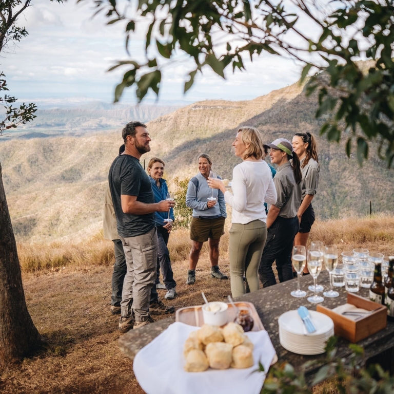 A group of hikers on the Scenic Rim Trail pause along the track to take in the sweeping views across a wide, green valley. They stand together on a natural lookout, surrounded by native bush and rugged terrain and enjoy afternoon drinks and snacks. The landscape stretches far into the distance, with rolling hills and layers of forest fading into the horizon. The hikers appear calm and reflective, enjoying a quiet moment of rest while appreciating the vast, open scenery and the beauty of the natural environment around them. © Tourism Australia