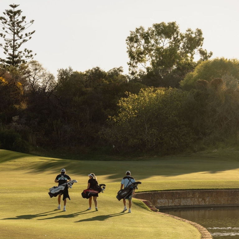 A group of golfers walks along a golden, sun-drenched fairway toward a green at Secret Harbour Golf Links in Secret Harbour, Western Australia. The course features a tranquil water hazard and lush coastal vegetation, creating a scenic backdrop for an afternoon round. © Tourism Australia