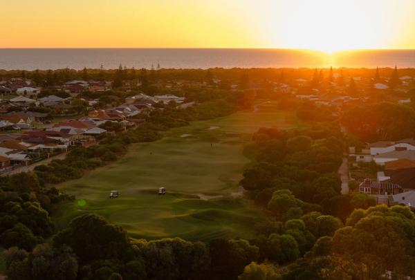 An aerial view of Secret Harbour Golf Links in Secret Harbour, Western Australia, during a spectacular golden sunset. The long fairway and undulating greens are bathed in warm light as the sun dips below the horizon of the Indian Ocean, with the surrounding coastal community nestled alongside the course. © Tourism Australia