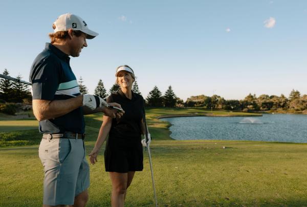 Two golfers share a smile and a conversation on the tee box at Secret Harbour Golf Links in Secret Harbour, Western Australia. The scenic hole features a large water hazard with a fountain, reflecting the clear blue sky of a late afternoon round south of Perth. © Tourism Australia
