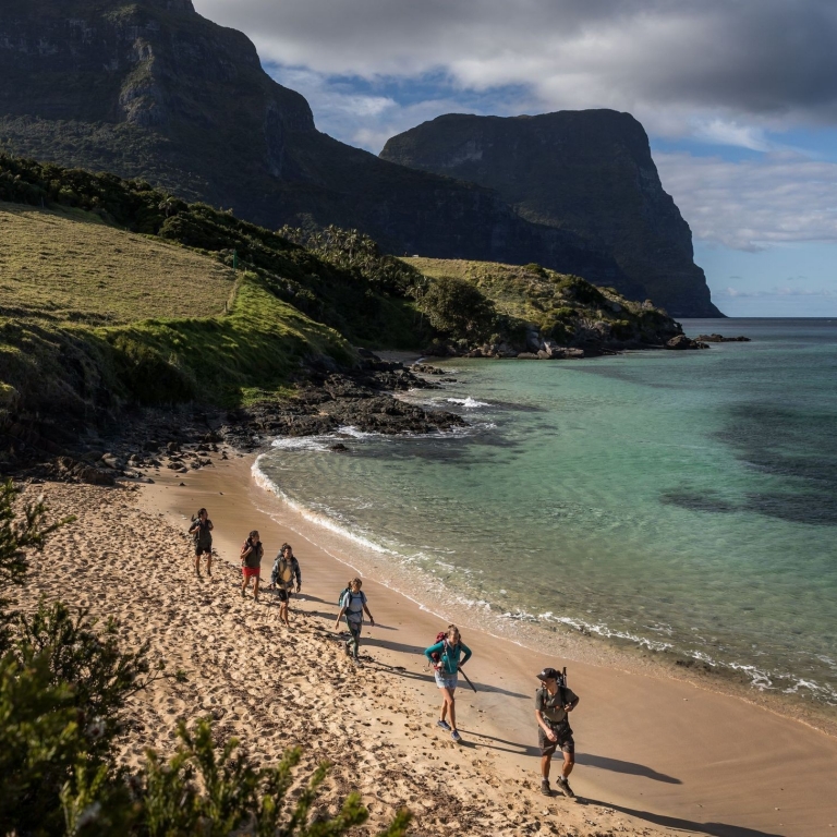 A group of hikers traipse across the golden sands of Lovers Beach on Lord Howe Island during the Seven Peaks Walk, New South Wales © Tourism Australia
