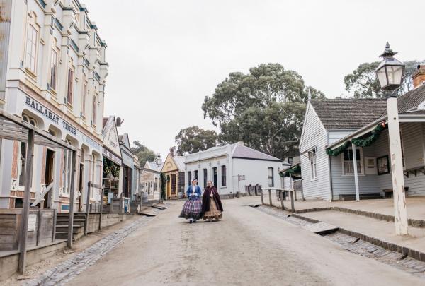 Two ladies dressed in period costumes walk down a street of old buildings at Sovereign Hill, Ballarat, Victoria © Tourism Australia