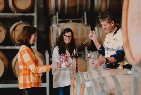 Inside a rustic cellar at Stanton and Killeen in the Murray River region, Victoria, a smiling winemaker uses a glass thief to pour wine from a barrel for two women, who hold up their glasses to enjoy a tasting experience. © Stanton & Killeen