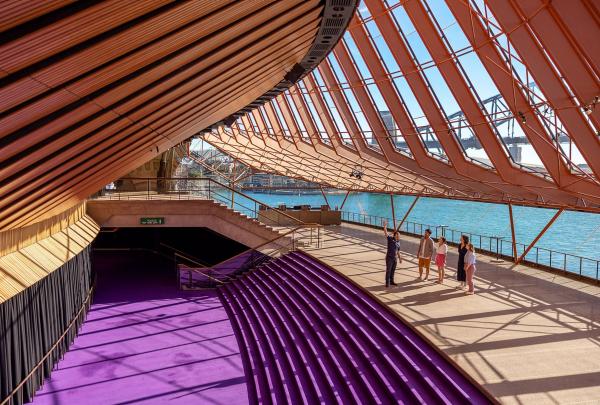 A diverse group of visitors immerse themselves in an architectural tour of the world-renowned Sydney Opera House. Against the backdrop of this iconic structure, they explore the numerous theatres and venues it houses, uncovering the profound design and engineering brilliance that earned it a coveted spot on the World Heritage List. The tour promises a deep dive into the heart of this cultural masterpiece, where history and innovation seamlessly converge. At times, the harbour and bridge can be seen in the background adding to this picturesque scene. © Tourism Australia