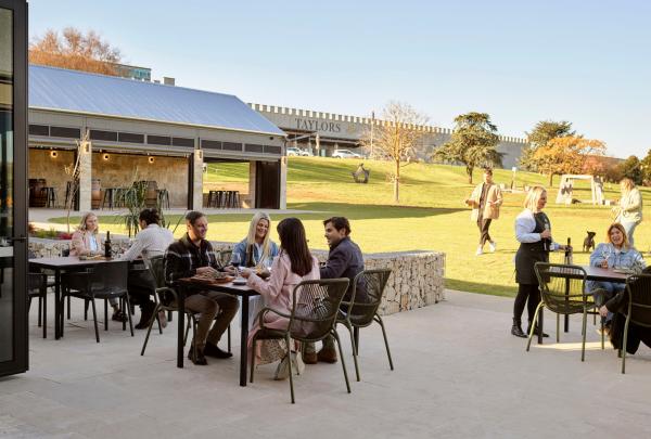 Guests enjoy an outdoor lunch and wine tasting on the sun-drenched terrace at Taylors Wines, South Australia. In the background, a lush green lawn leads toward the iconic winery building set against a clear, bright sky. © Taylors Wines