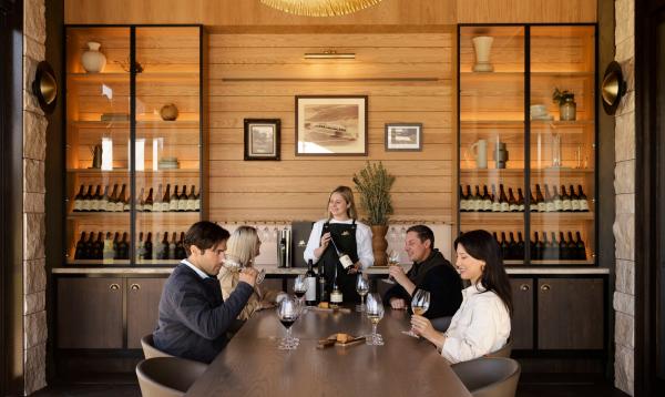 Four guests enjoy a guided tasting at Taylors Wines, South Australia. A server presents a bottle at a long timber table, while guests sip wine beneath a striking, oversized gold pendant light in a modern cellar. © Taylors Wines