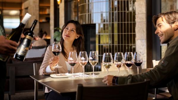 A woman in a beige blazer and a man enjoy a wine flight at Taylors Wines, South Australia. A server presents bottles as they taste seven different wines at a table inside the sleek, modern cellar door. © Taylors Wines