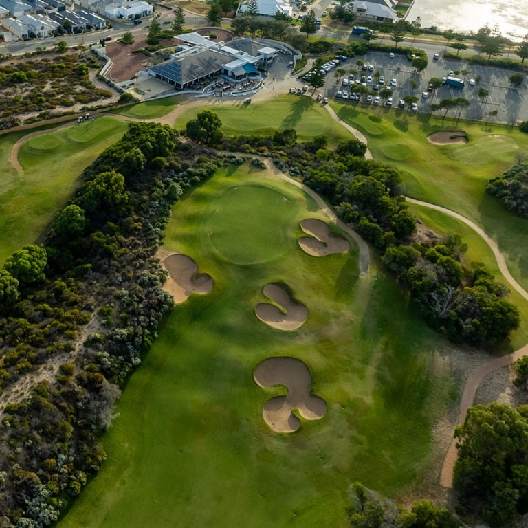 An aerial view of The Cut Golf Course in Mandurah, Western Australia, showcasing its dramatic links-style layout nestled along the coastline. The bright green fairways wind through rugged dunes and native scrub, bordering the white sands and turquoise waters of the Indian Ocean. © The Cut Golf Course