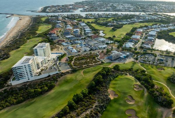 An aerial view of The Cut Golf Course in Mandurah, Western Australia, showcasing its dramatic links-style layout nestled along the coastline. The bright green fairways wind through rugged dunes and native scrub, bordering the white sands and turquoise waters of the Indian Ocean. © The Cut Golf Course