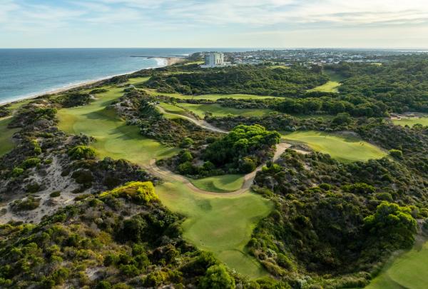 An aerial view of The Cut Golf Course in Mandurah, Western Australia, showcasing its dramatic links-style layout nestled along the coastline. The bright green fairways wind through rugged dunes and native scrub, bordering the white sands and turquoise waters of the Indian Ocean. © The Cut Golf Course
