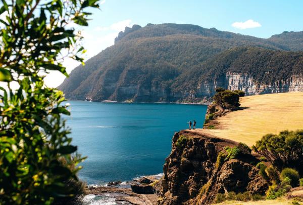A pair hike along a rugged cliff edge on Maria Island during The Maria Island Walk. The sun shines brightly overhead, casting warm light on the rocky landscape. Below, the dramatic cliffs drop sharply to reveal the deep blue ocean stretching out to the horizon, creating a stunning and peaceful scene. © The Maria Island Walk & Great Walks of Australia