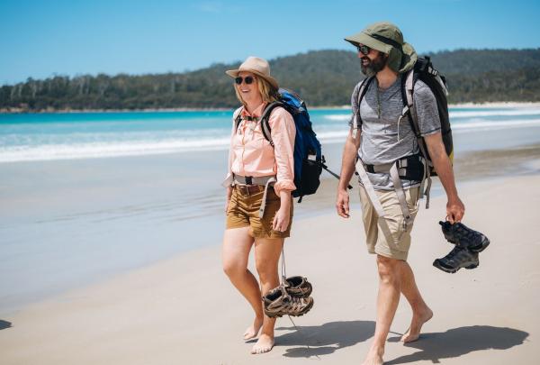 Two people walk barefoot along a white sandy beach on Maria Island, carrying their boots in hand on The Maria Island Walk, Tasmania © Tourism Australia