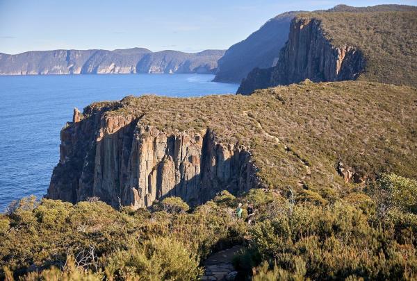 A general view of a rugged outcrop at Cape Hauy. The sun streams through dramatic rock formations, casting shadows across the cliffs. The jagged rocks rise sharply from the sea below, showcasing the raw beauty of the coastline, Three Capes Signature Walk, Tasmanian Walking Co, Tasmania © Tourism Australia