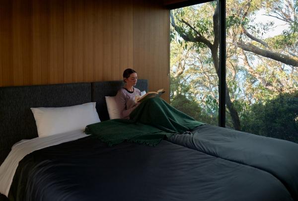 A person relaxes on their comfortable bed inside a cosy room at Cape Pillar Lodge, Tasmanian Walking Co, Tasmania © Tourism Australia