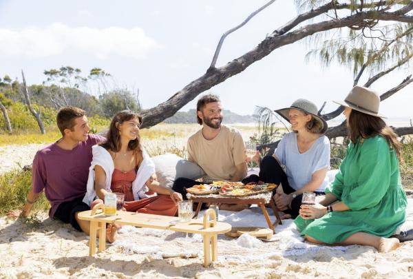 Group of talent on picinic rug at beach enjoying seafood and cheese on the Indigenous Tour, Yura Tours, Minjerribah/North Stradbroke Island, Queensland © Tourism Australia